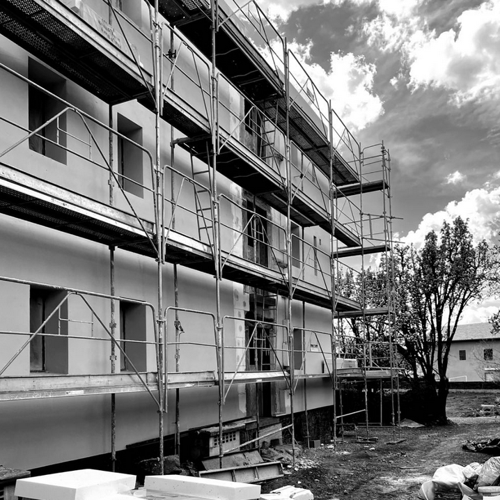 Façade d’un bâtiment en cours de chantier, entourée d’échafaudages sur plusieurs niveaux. Des panneaux d’isolation thermique par l’extérieur sont en cours d’application sur les murs, avec des zones partiellement enduites. Le ciel nuageux et le terrain en contrebas complètent cette scène de transformation, témoignant d’une étape importante de la rénovation énergétique du projet Le Jardin.