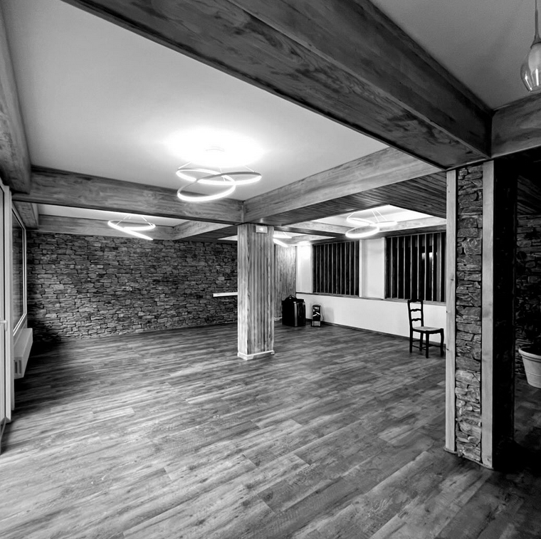 Salle de petit déjeuner du projet Le Jardin. Espace vaste et lumineux, habillé de bois au sol et en poutres apparentes, murs en pierre et grandes fenêtres verticales apportant de la clarté. Des luminaires circulaires suspendus apportent une touche contemporaine dans une ambiance à la fois chaleureuse et minérale.