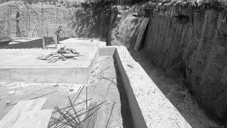 Vue du chantier de terrassement du projet à Targasonne. Une tranchée profonde a été creusée dans le terrain, révélant les strates naturelles du sol. La dalle de béton est en cours de réalisation avec ferraillage apparent au premier plan. Quelques outils et matériaux sont disposés sur le chantier, sous un ciel dégagé, entouré d’arbres en arrière-plan.
